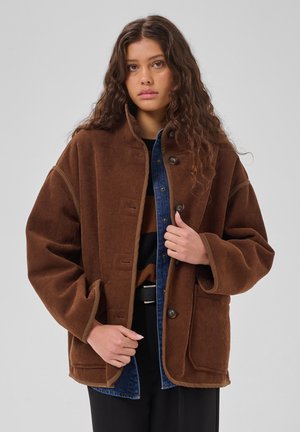 Jeune femme aux longs cheveux bouclés portant une veste en velours côtelé marron par-dessus une chemise en jean bleue et un pull à rayures sur un fond uni.