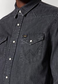 Tummanharmaa farkkupaita, jossa on klassinen kaulus, nepparit, kaksi rintataskua, joissa on sahalaitakuviota, ja pieni logo.