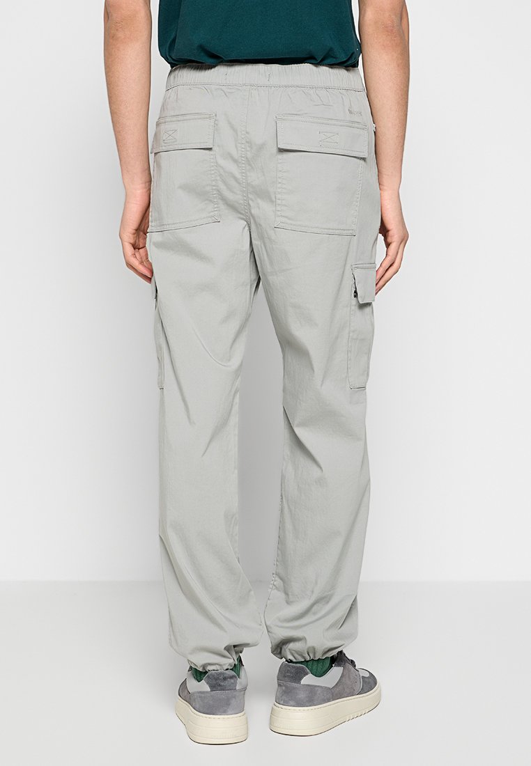 Hellgraue Cargo-Hosen mit elastischem Bund, zwei Gesäßtaschen und zusätzlichem seitlichen Taschendetail, kombiniert mit grauen Sneakern.