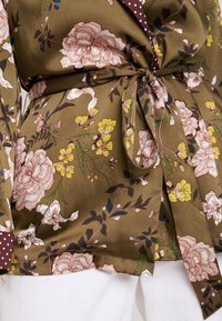 Blouse cache-cœur florale vert olive avec des fleurs roses et jaunes nouée à la taille, avec col et poignets à pois marron, portée avec un pantalon blanc.