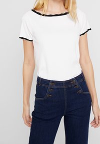 Mujer vestida con una blusa blanca de manga corta con ribetes negros y jeans oscuros de tiro alto con seis botones en los bolsillos delanteros.