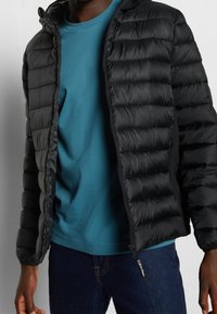 Sort pufferjakke med vandret quiltning og lynlås, båret over en teal crew neck t-shirt. Har kompakt design og elastiske manchetter.