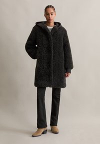 Zwarte bontjas met een textuurrijke, fuzzy buitenkant, voorzien van een capuchon, zijzakken en een ruime silhoeet. Gepaard met zwarte broek en beige hakken.