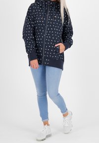 Kvinna i en marinblå zip-up hoodie med vita fågelmönster, ljusblå jeans och vita sneakers, stående mot en vit bakgrund.