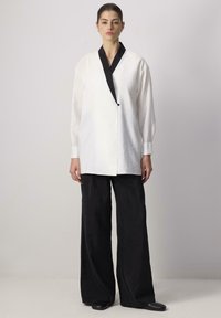 White long-sleeve shirt with black collar and button detail, paired with loose black pants made from textured fabric, worn close-fitting shoes.