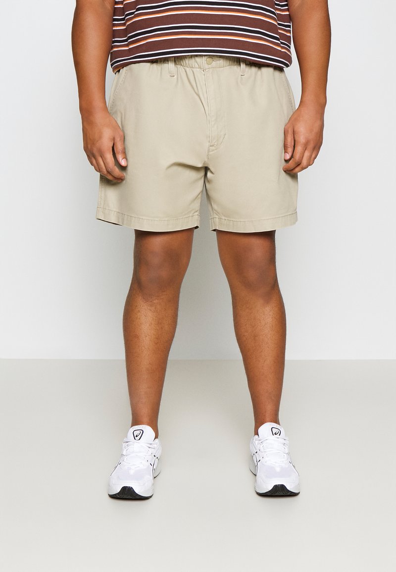 Khakifarbenen Baumwollshorts mit einem elastischen Bund und umgeschlagenem Saum, kombiniert mit einem braun-gestreiften Kurzarmhemd und weißen Sneakers.
