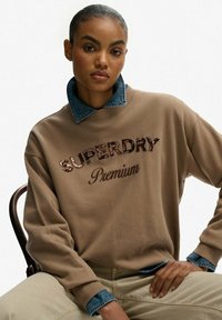 Beżowa bluza ze dżinsowym kołnierzem, z wypukłym napisem "SUPERDRY" oraz "Premium" w kursywie, lekko luźny krój, miękka tekstura.