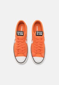 Pomarańczowe trampki z płótna z białymi gumowymi czubkami i czarnymi podeszwami. Zawierają czarną naszywkę z logo "Converse All Star". Zapięcie na sznurówki.