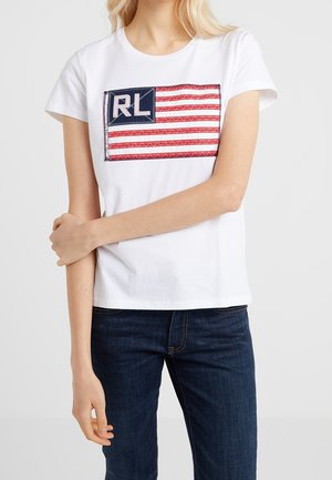 T-shirt en coton blanc avec un patch sur le devant affichant un design de drapeau américain rouge et bleu. Le t-shirt a un col rond classique et des manches courtes.
