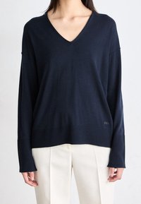 Kobieta w ciemnoniebieskim swetrze z dekoltem w serek i długim rękawem, z prążkowanymi mankietami i dolnym brzegiem, w połączeniu z wysokim stanem kremowymi spodniami plisowanymi.