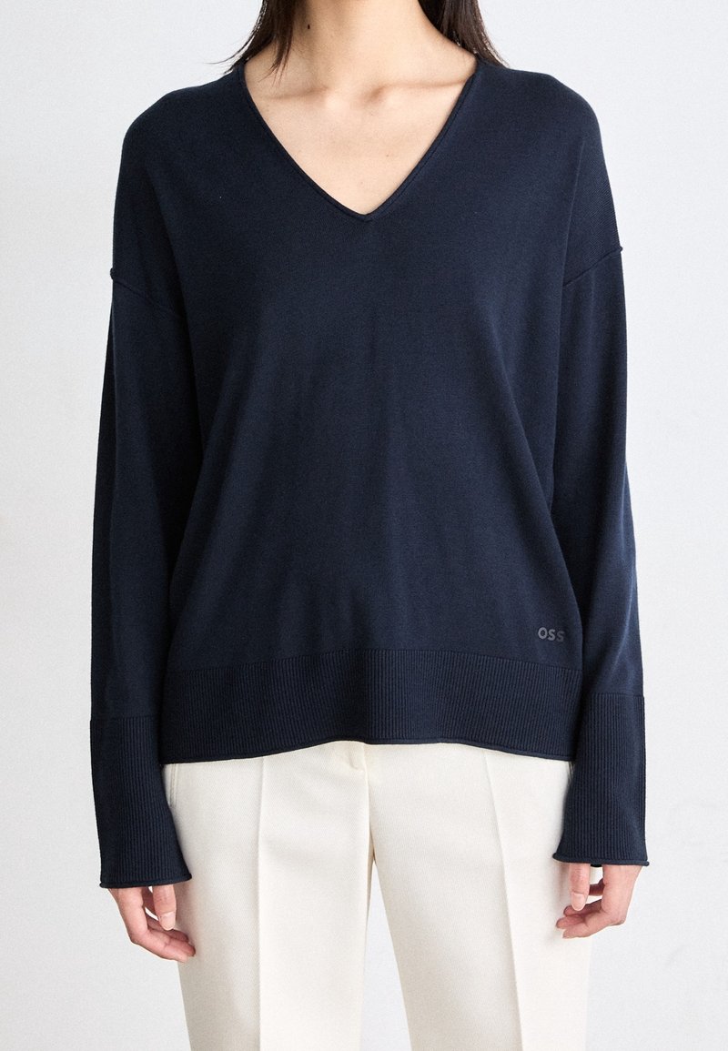 Kobieta w ciemnoniebieskim swetrze z dekoltem w serek i długim rękawem, z prążkowanymi mankietami i dolnym brzegiem, w połączeniu z wysokim stanem kremowymi spodniami plisowanymi.