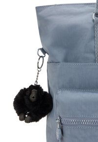 Musta pörröinen gorillan avaimenperä roikkuu sinisen kankaisen laukun edessä olevan vetoketjullisen taskun ja hihnan päällä valkoista taustaa vasten.