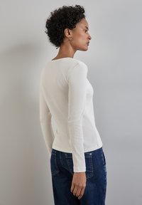 Weißes, geripptes Langarmshirt mit rundem Halsausschnitt, kombiniert mit dunkelblauen Jeans. Der Stoff wirkt strukturiert, figurbetont und glatt.