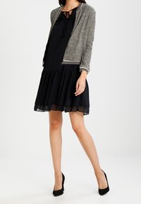 Grå stickad bomberjacka över en svart klänning med utsvängd kjol. Klänningen har en knytkrage och ett genomskinligt fåll. Svarta högklackade skor.