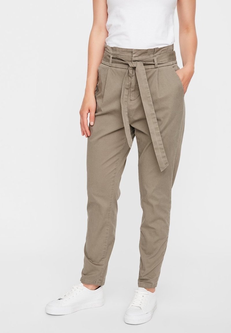 Personne portant un pantalon beige taille haute paperbag avec ceinture nouée devant, des baskets blanches et un débardeur blanc, debout avec une main dans la poche.