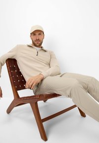 Beige Zip-up-Sweatshirt und passende Hose, getragen von einer Person, die auf einem Stuhl aus Holz mit geflochtener Sitzfläche sitzt, vor einem weißen Hintergrund.