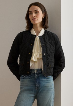 Kvinde iført en sort quiltet jakke over en hvid bluse med sløjfe og lyseblå jeans, stående foran en neutral væg.
