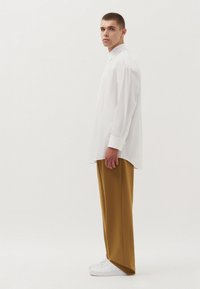 Witte lange mouwen shirt met een kraag, gecombineerd met mosterdkleurige broek met brede pijpen en witte sneakers, gedragen door een mannelijke model die zijwaarts kijkt.