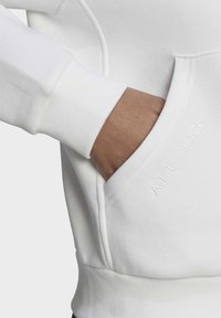 Haut à capuche en coton blanc avec des poignets côtelés et une poche kangourou. Présente une broderie subtile "ALL SZN" sur la poche, soulignant un design minimaliste.