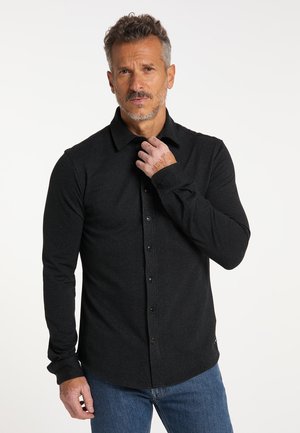 Mann mit salt-and-pepper-Haaren, der ein schwarzes Hemd mit Knöpfen und blaue Jeans trägt, richtet mit seiner rechten Hand seinen Kragen.