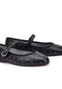 Bailarinas de cuero negro con punta afilada, adornadas con tachuelas plateadas y una correa en el tobillo decorativa con una hebilla metálica.