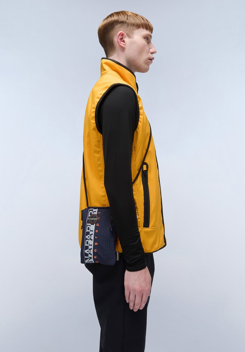 Gilet isolante giallo con bordi neri, dotato di tasche laterali. Indossato sopra una maglietta nera a maniche lunghe, con una borsa blu navy attaccata.