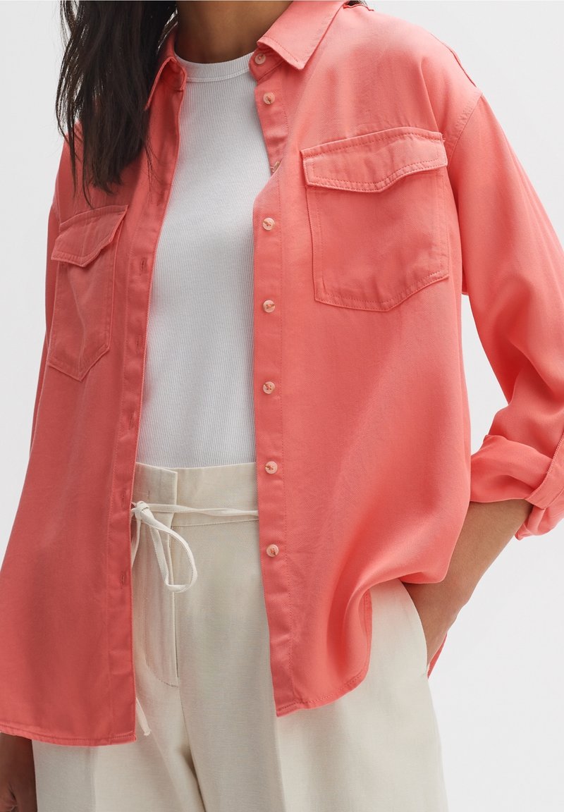 Femme portant une chemise corail à boutons par-dessus un débardeur blanc côtelé et un pantalon crème taille haute avec cordon, une main dans la poche.