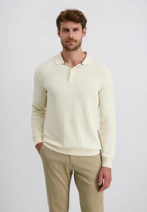 Cremfarbener gestrickter Poloshirt mit langen Ärmeln, gerippter Textur und flacher Kragen. Kombiniert mit beigen Hosen, Hände in den Taschen.