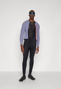 Conjunto atlético para hombres que incluye una chaqueta morada de manga corta con cremallera rosa, una camiseta de malla negra y leggings ajustados negros.