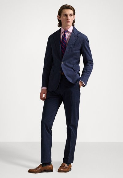 Ναυτικό βαμβακερό κοστούμι με τακάριστη εφαρμογή, χαρακτηριστικό του οποίου είναι το γιακάκι notch, η τσέπη στο στήθος, συνδυασμένο με ροζ πουκάμισο και ριγέ γραβάτα. Καφέ λόφερς.