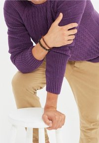 Violetti ribbineulottu paita, jossa on teksturoitu neulos, puettu beigejen housujen päälle. Kädet lepäävät valkoisella jakkaralla, toisessa ranteessa helmikoristeinen rannekoru.