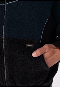 Fleecejacka i mörk marinblå och svart, med dragkedja fram, strukturerad tyg och en silverfärgad metall-logotyp på fickan.