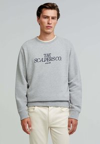 Gray sweatshirt made of cotton with a crew neck and long sleeves. Features navy blue logo text on the front. Worn with light-colored pants.