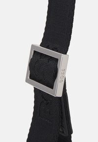 Juodas tinklelis dirželis su kvadratine metaline sagtimi. Sagtis yra sidabrinės spalvos su iškilmingu prekės ženklu. Dirželis turi tekstūruotą paviršių.