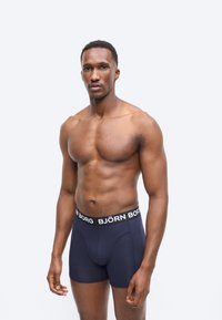 Μυώδης άνδρας με κοντά μαλλιά, φορώντας σκούρα μπλε μποξεράκια Björn Borg, στέκεται μπροστά σε απλό ανοιχτό φόντο.