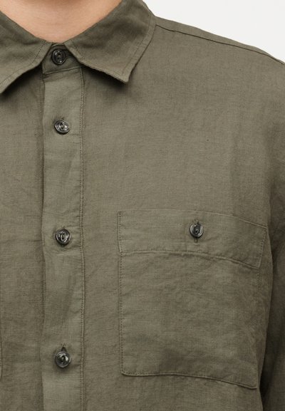 Πράσινο-ελαιί πουκάμισο με κουμπιά και μπροστινή τσέπη, εμφανίζεται στο πάνω μέρος του κορμού ενός ατόμου.