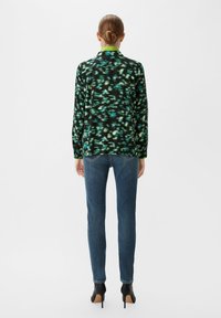 Long-sleeve shirt in black with green and blue abstract pattern, paired with fitted blue jeans and black high-heeled shoes.
