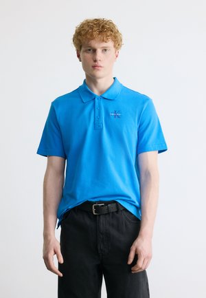 Fiatal férfi göndör hajjal, kék Calvin Klein galléros pólót és fekete nadrágot visel, sima fehér háttér előtt.