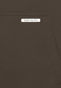 Tissu marron avec une texture lisse, featuring une petite étiquette métallique avec l'inscription "bazir republiq," détail de poche cousue sur le côté.