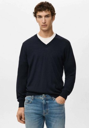 Junger Mann mit lockigem Haar, der einen schwarzen V-Ausschnitt-Pullover über einem weißen Hemd und blauen Jeans trägt, mit einer Hand in der Tasche, steht vor einem schlichten Hintergrund.