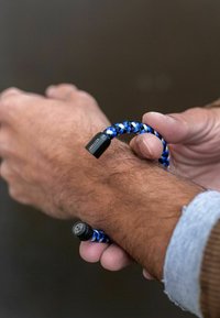 Mėlynas ir baltas pynėtas apyrankė su juodu furnitūru, kuriame viename gale yra lotoso dizainas, uždėta ant riešo.