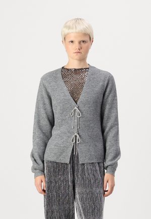 Cárdigan gris con acolchado y de manga larga, con escote en V y cierre con cordón. Debajo, una camiseta de malla negra con hilos metálicos.