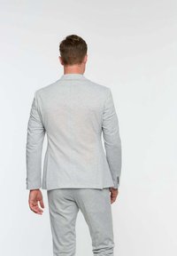 Mężczyzna stojący plecami, ubrany w jasnoszary garnitur z długimi rękawami i dopasowane spodnie na jednolitym białym tle.