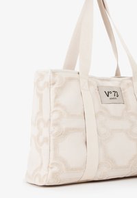 Sac en toile beige avec un motif géométrique texturé, doté de côtés rembourrés et de deux poignées en tissu pour un transport facile.