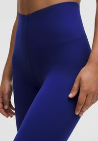 Leggings taille haute, ajustés, en tissu bleu foncé. Texture lisse avec un léger brillant. Détails de couture visibles près de la taille.