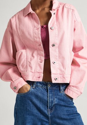 Persona vistiendo una chaqueta vaquera corta de color rosa claro sobre un top corto texturizado magenta, combinados con vaqueros azules de tiro alto y con las manos en los bolsillos.