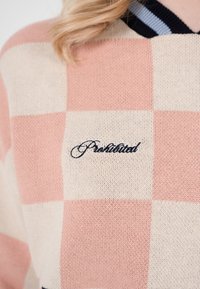 Prim-plan al unui pulover în carouri, roz și crem, cu cuvântul „Practical” brodat în scris negru pe zona pieptului.