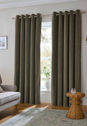 Dicke dunkelgrüne Vorhänge hängen an einer silbernen Stange über einem Fenster mit einer weißen Vase und Blumen auf der Fensterbank, neben einem grauen Sessel und einem Korb-Beistelltisch.