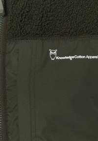 Fleecefodrad jacka med en jämn, mörkgrön utsida. Funktioner inkluderar dragkedjestängning och en vit logotyp av KnowledgeCotton Apparel.