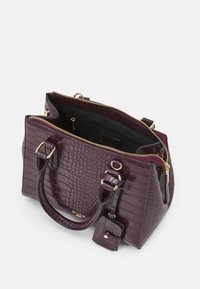 Torebka w kolorze purpurowym z wzorem krokodyla, z złotymi elementami, podwójnymi uchwytami i zapięciem na zamek. Wnętrze ma czarne wykończenie oraz kieszeń.